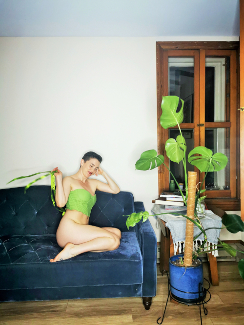 A white artist with short brown hair tosses the green ribbons of their corset in the air sitting next to a green plant
